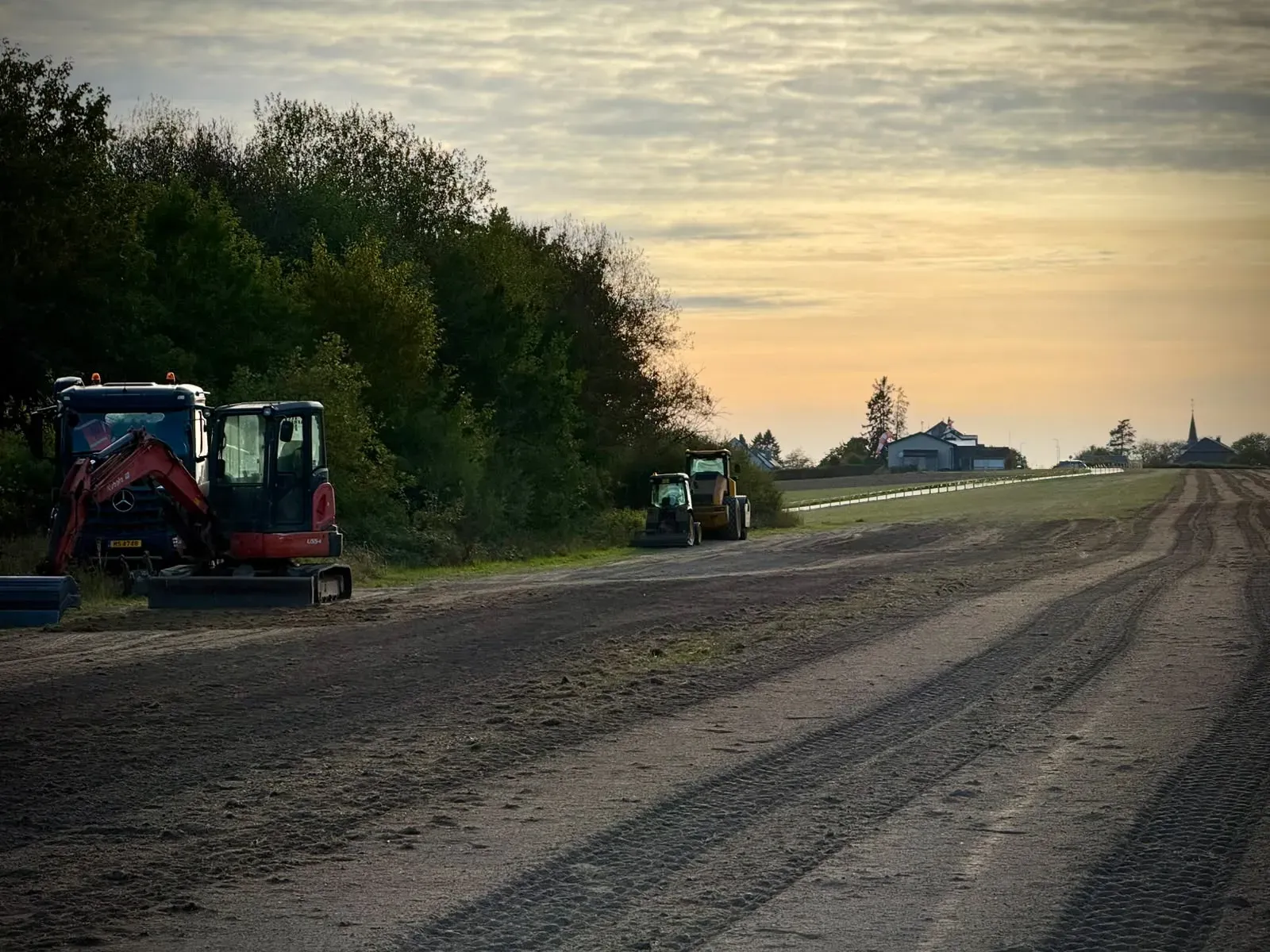 Machines parked along the treeline beside the runway at dusk, with the village and its church spire on the horizon