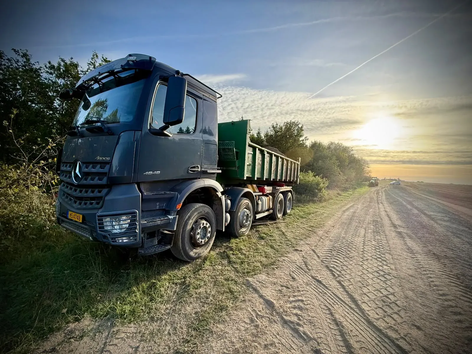 A heavy truck with a loaded container parked on the grass beside the runway, the setting sun beyond